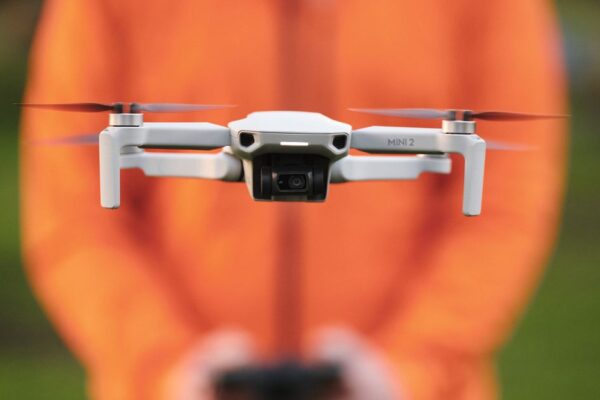 DJI afirma que su decisión de permitir que los drones vuelen en áreas peligrosas no es política. A drone faces the camera, in flight, with a blurred person in the background shoulders down in an orange puffy jacket controlling that drone, serving as an orange background.